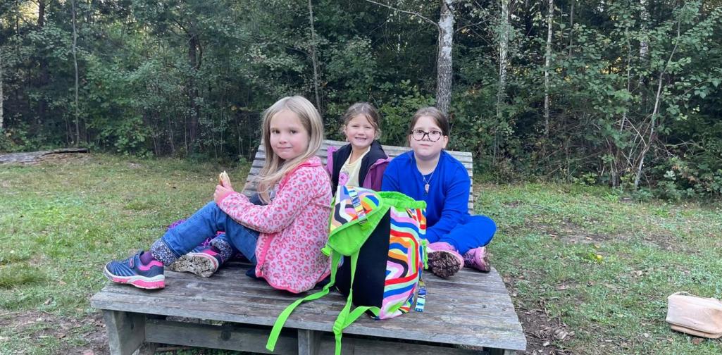 VS Amras Drei Kinder sitzen während der Jausenpause gemeinsam auf einer Holzplattform im Wald.