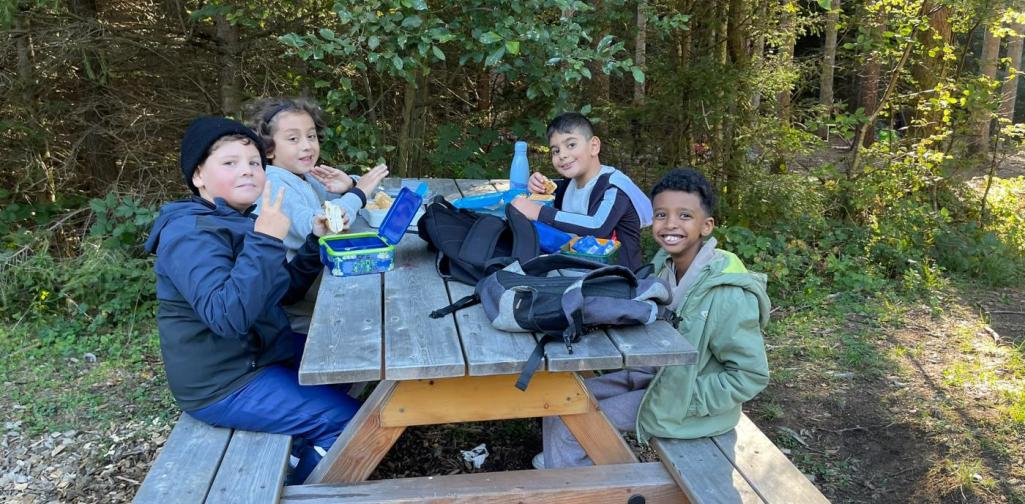 VS Amras Vier Kinder sitzen gemeinsam bei einer Jausenpause an einem Holztisch im Wald.