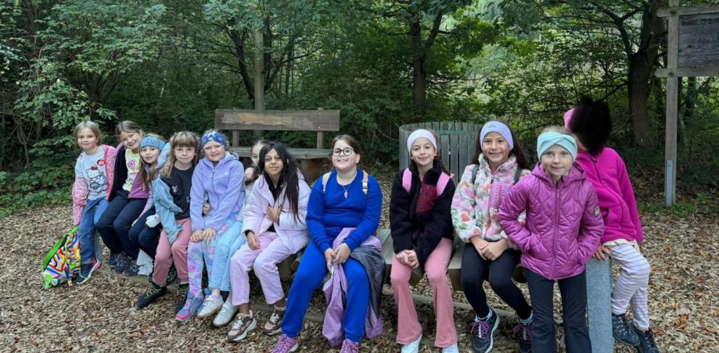 VS Amras Kinder der 2A und 2B sitzen gemeinsam auf einer Bank im Wald und posieren für ein Gruppenfoto.