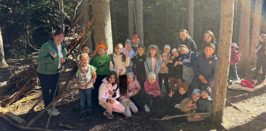 VS Amras Kinder und Lehrpersonen stehen gemeinsam vor einem selbstgebauten Unterschlupf im Wald.